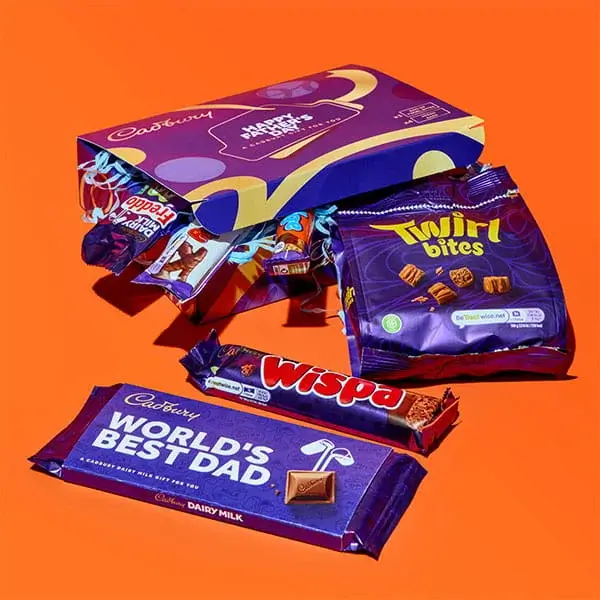 A selection of Cadbury chocolate gifts including a Worlds Best Dad Dairy Milk bar, Wispa bar, Twirl Bites pack and assorted chocolates displayed on a bright orange background.